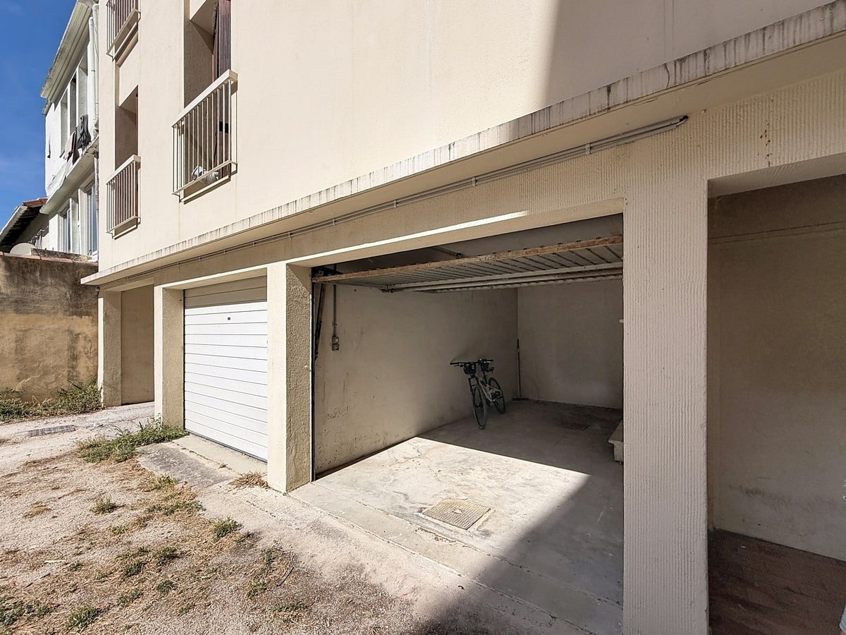 Garage individuel en rez-de-chaussée d'un immeuble aux murs enduits beige, avec porte sectionnelle relevable en métal blanc partiellement ouverte, un vélo visible à l'intérieur. Le sol est en béton brut et le bâtiment présente des balcons en étages supérieurs.