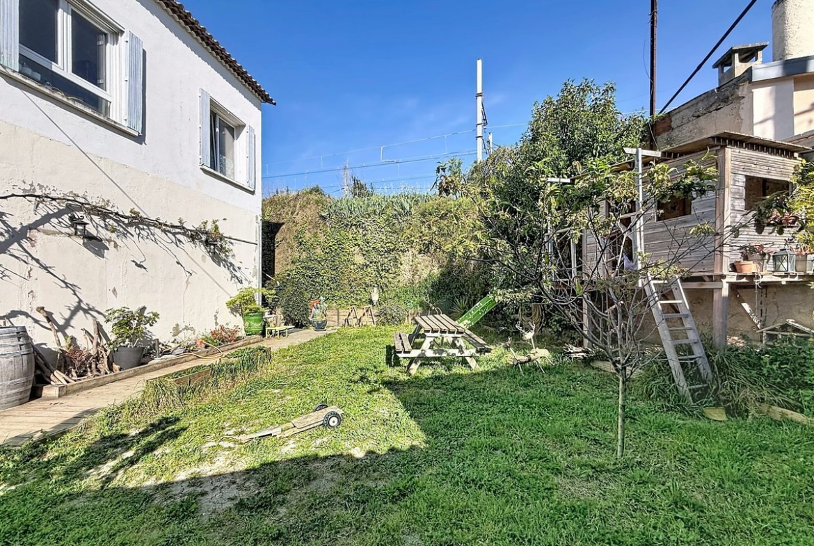 Vue du jardin depuis l'angle de la façade blanche de la maison, montrant une pelouse verte avec des arbres et arbustes en arrière-plan et une cabane en bois sur pilotis avec escalier sur la droite. Des pots de fleurs, un tonneau en bois et divers objets de jardin sont disposés contre le mur de la maison sous un ciel bleu dégagé.