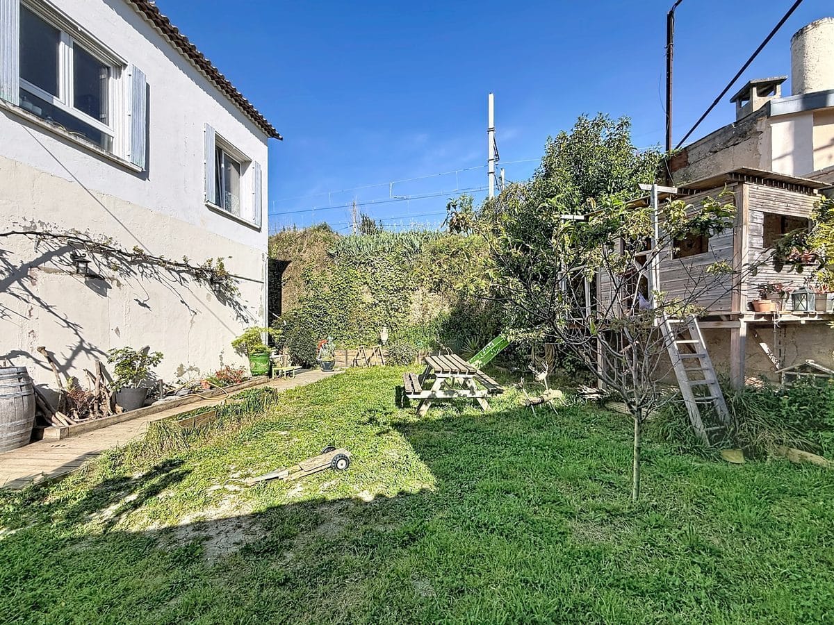Vue du jardin depuis l'angle de la façade blanche de la maison, montrant une pelouse verte avec des arbres et arbustes en arrière-plan et une cabane en bois sur pilotis avec escalier sur la droite. Des pots de fleurs, un tonneau en bois et divers objets de jardin sont disposés contre le mur de la maison sous un ciel bleu dégagé.