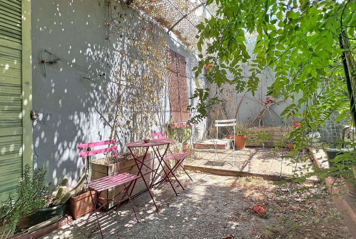 Jardin en cour intérieure avec une petite terrasse en gravier et dalles, meublée d'une table et deux chaises pliantes en métal rose fuchsia, entourée de plantations variées dont des cactus et des plantes grimpantes. Les murs mitoyens peints en gris sont recouverts de filets et de végétation, un arbre à feuilles vertes apporte de l'ombre à l'espace.