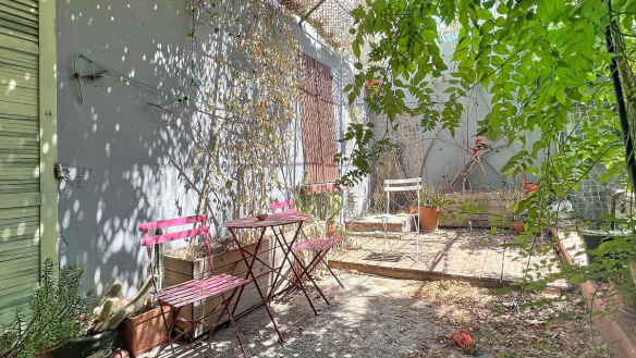 Jardin en cour intérieure avec une petite terrasse en gravier et dalles, meublée d'une table et deux chaises pliantes en métal rose fuchsia, entourée de plantations variées dont des cactus et des plantes grimpantes. Les murs mitoyens peints en gris sont recouverts de filets et de végétation, un arbre à feuilles vertes apporte de l'ombre à l'espace.