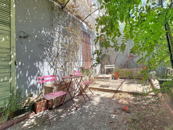 Jardin en cour intérieure avec une petite terrasse en gravier et dalles, meublée d'une table et deux chaises pliantes en métal rose fuchsia, entourée de plantations variées dont des cactus et des plantes grimpantes. Les murs mitoyens peints en gris sont recouverts de filets et de végétation, un arbre à feuilles vertes apporte de l'ombre à l'espace.