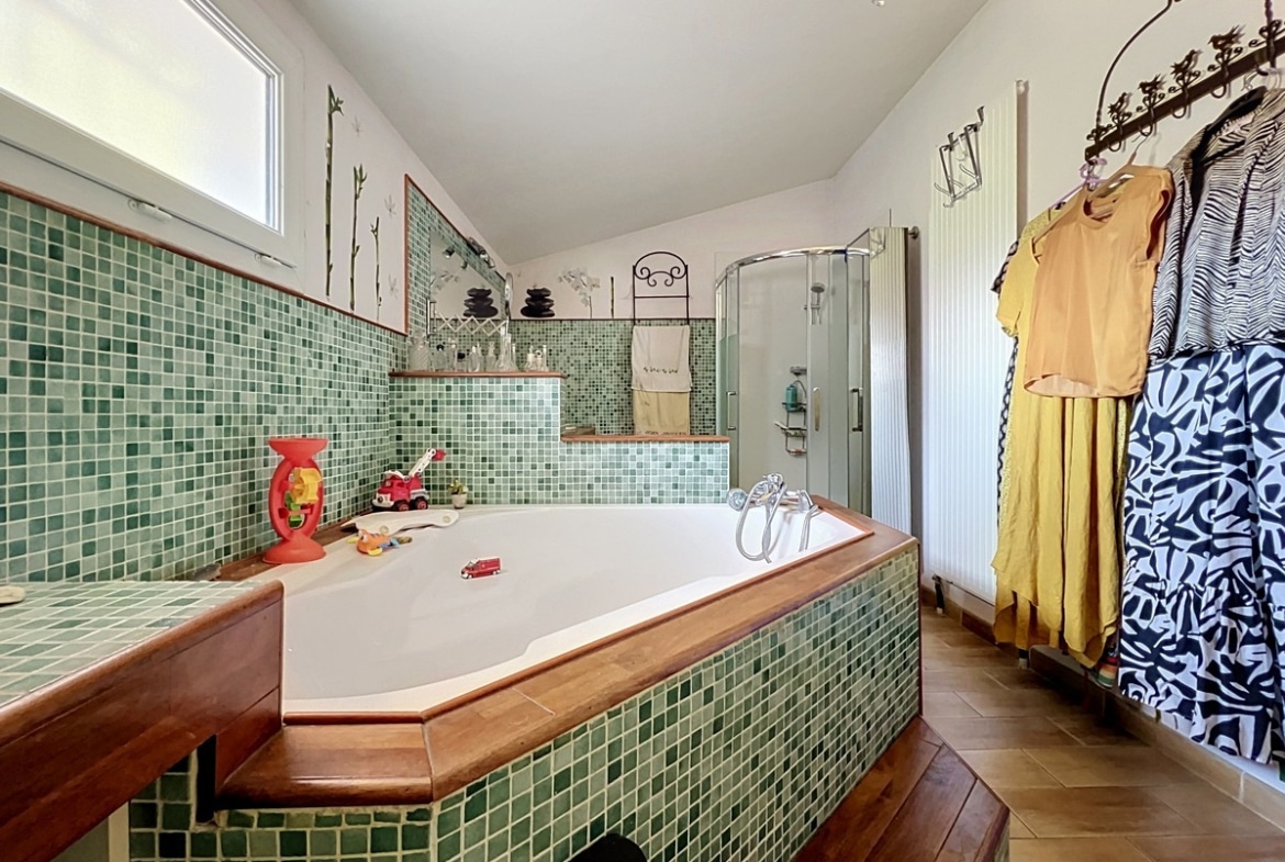 Salle de bain avec baignoire balnéo hexagonale habillée de mosaïque verte et encadrée de bois exotique, posée sur un podium en carrelage bois. Cabine de douche en verre dépoli dans l'angle, murs partiellement carrelés de mosaïque verte avec décoration florale peinte sur la partie haute blanche, fenêtre en double vitrage côté gauche.