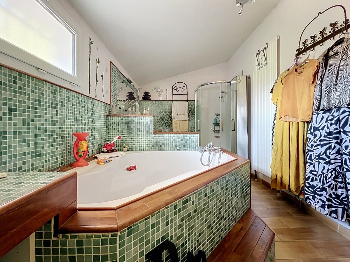 Salle de bain avec baignoire balnéo hexagonale habillée de mosaïque verte et encadrée de bois exotique, posée sur un podium en carrelage bois. Cabine de douche en verre dépoli dans l'angle, murs partiellement carrelés de mosaïque verte avec décoration florale peinte sur la partie haute blanche, fenêtre en double vitrage côté gauche.