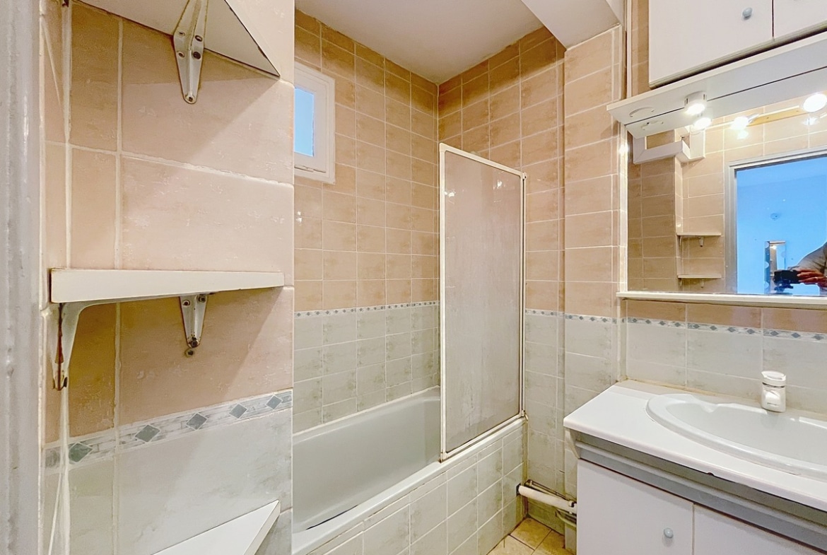Salle de bain carrelée du sol au plafond avec faïence beige rosé et frise décorative en losanges gris-bleu, équipée d'une baignoire encastrée avec pare-baignoire en verre coulissant à cadre aluminium. Un meuble vasque blanc avec miroir éclairé et rangements hauts est installé sur le mur de droite, et une petite fenêtre en hauteur assure la ventilation naturelle.