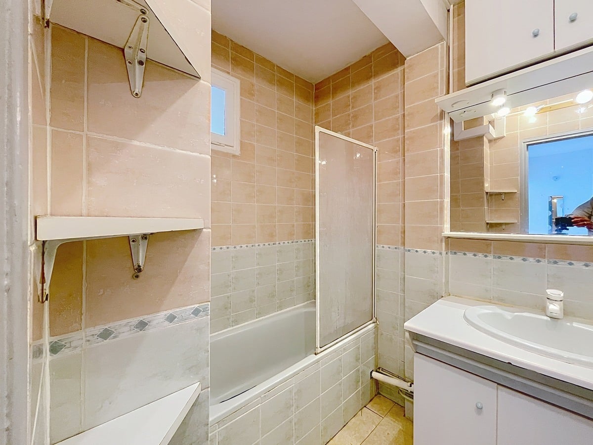 Salle de bain carrelée du sol au plafond avec faïence beige rosé et frise décorative en losanges gris-bleu, équipée d'une baignoire encastrée avec pare-baignoire en verre coulissant à cadre aluminium. Un meuble vasque blanc avec miroir éclairé et rangements hauts est installé sur le mur de droite, et une petite fenêtre en hauteur assure la ventilation naturelle.