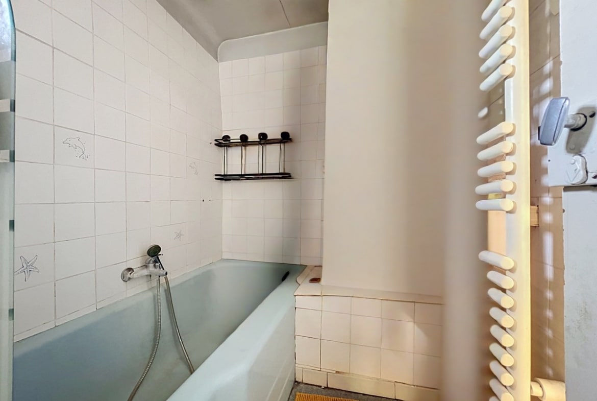Salle de bain carrelée de faïence blanche avec une baignoire encastrée de couleur vert d'eau équipée d'un flexible de douche, et un sèche-serviettes à barreaux blancs fixé au mur. Des étagères métalliques noires sont posées sur le carrelage au-dessus de la baignoire, le sol est recouvert d'un tapis de bain beige.