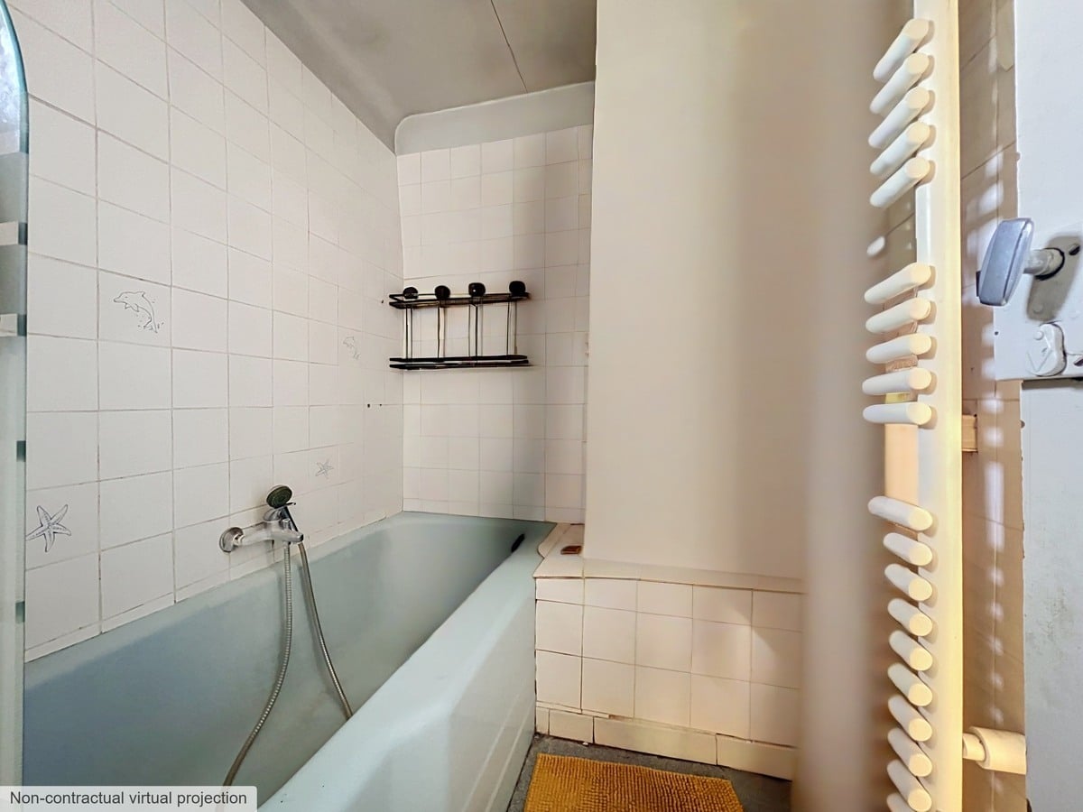 Salle de bain carrelée de faïence blanche avec une baignoire encastrée de couleur vert d'eau équipée d'un flexible de douche, et un sèche-serviettes à barreaux blancs fixé au mur. Des étagères métalliques noires sont posées sur le carrelage au-dessus de la baignoire, le sol est recouvert d'un tapis de bain beige.