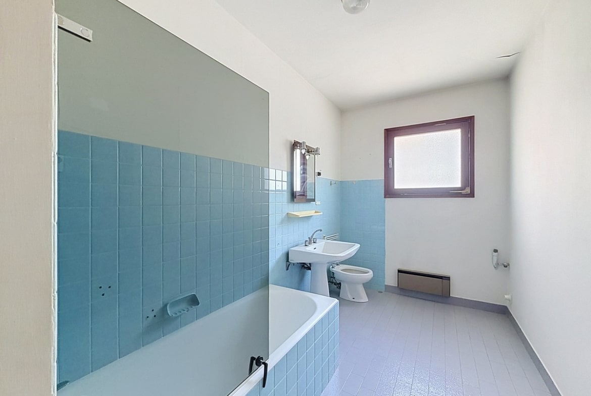 Salle de bain avec carrelage mural en faïence bleue posé à mi-hauteur, comprenant une baignoire encastrée bleue, un lavabo sur colonne blanc, un bidet et des WC blancs. Le sol est recouvert d'un revêtement souple gris-mauve et une fenêtre avec encadrement violet apporte de la lumière naturelle.