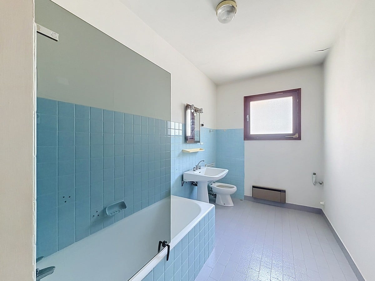 Salle de bain avec carrelage mural en faïence bleue posé à mi-hauteur, comprenant une baignoire encastrée bleue, un lavabo sur colonne blanc, un bidet et des WC blancs. Le sol est recouvert d'un revêtement souple gris-mauve et une fenêtre avec encadrement violet apporte de la lumière naturelle.