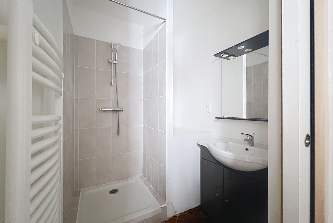Salle de bain avec cabine de douche carrelée en grand format gris clair et meuble vasque noir avec miroir rétroéclairé. Sol en tomettes hexagonales en terre cuite et sèche-serviettes à droite.