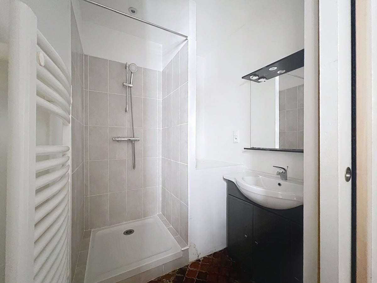 Salle de bain avec cabine de douche carrelée en grand format gris clair et meuble vasque noir avec miroir rétroéclairé. Sol en tomettes hexagonales en terre cuite et sèche-serviettes à droite.