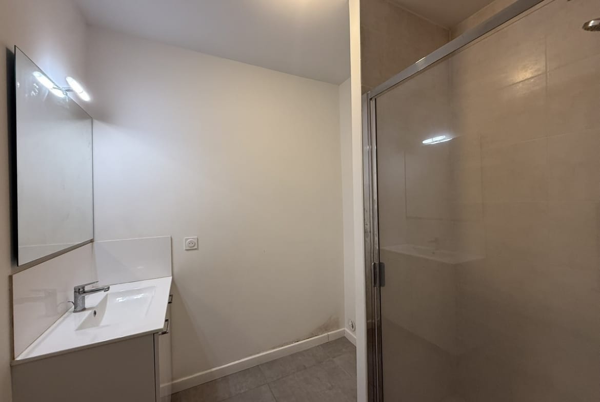 Salle de bain avec meuble vasque blanc suspendu surmonté d'un miroir avec bandeau lumineux LED, et cabine de douche fermée par une paroi en verre transparent à droite. Le sol est recouvert de carrelage gris clair et les murs sont peints en blanc.