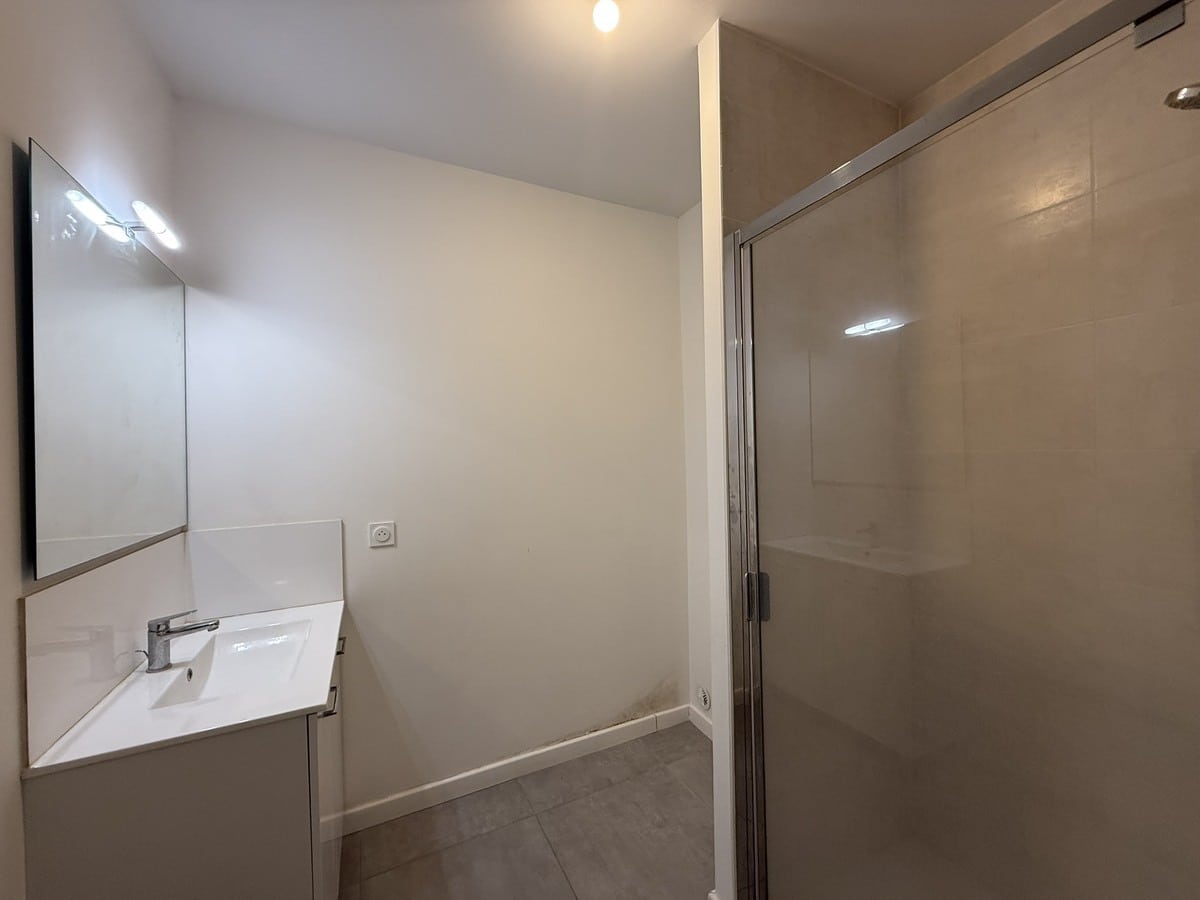 Salle de bain avec meuble vasque blanc suspendu surmonté d'un miroir avec bandeau lumineux LED, et cabine de douche fermée par une paroi en verre transparent à droite. Le sol est recouvert de carrelage gris clair et les murs sont peints en blanc.