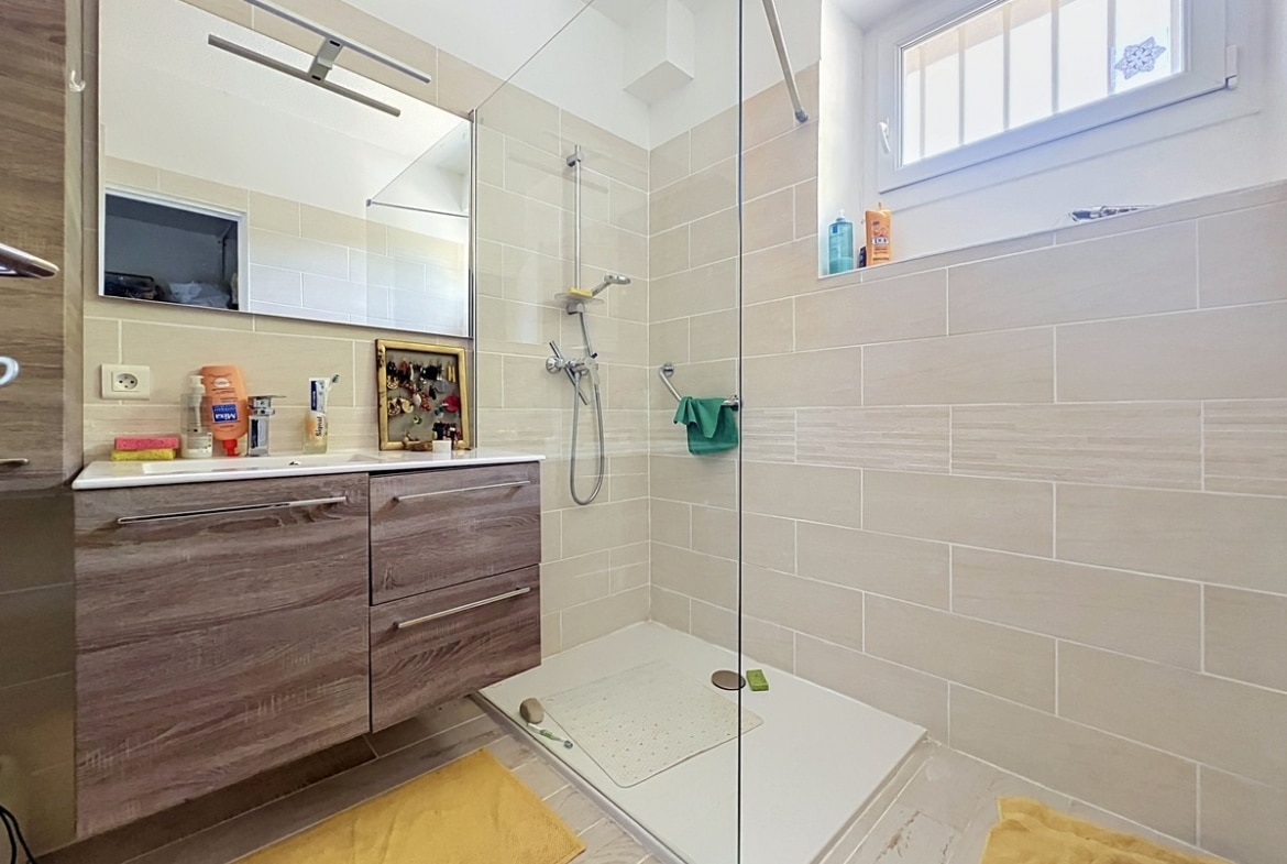 Salle de bain avec paroi de douche en verre transparent, meuble vasque suspendu en bois foncé et carrelage mural en grès beige à format rectangulaire. Fenêtre à double vitrage avec grille décorative apportant une lumière naturelle directe sur la vasque blanche encastrée.