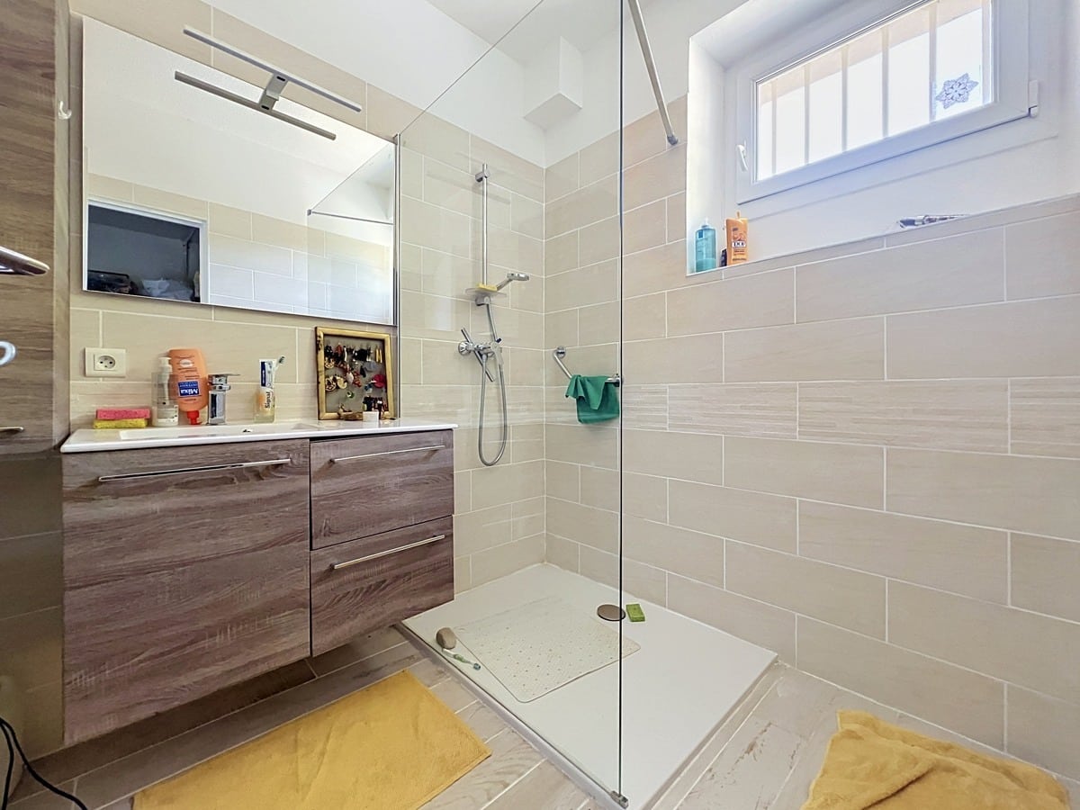 Salle de bain avec paroi de douche en verre transparent, meuble vasque suspendu en bois foncé et carrelage mural en grès beige à format rectangulaire. Fenêtre à double vitrage avec grille décorative apportant une lumière naturelle directe sur la vasque blanche encastrée.