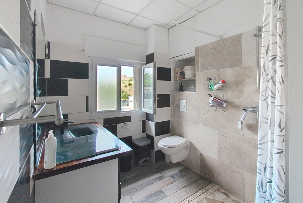 Salle d'eau avec carrelage mural en damier noir et blanc, sol en dalles imitation bois gris et ancien carrelage en terre cuite, équipée d'un WC suspendu blanc, d'une douche à l'italienne avec rideau à motifs végétaux et d'un meuble vasque noir à plan de travail sombre. Une fenêtre à battants apporte de la lumière naturelle et donne vue sur des arbres.
