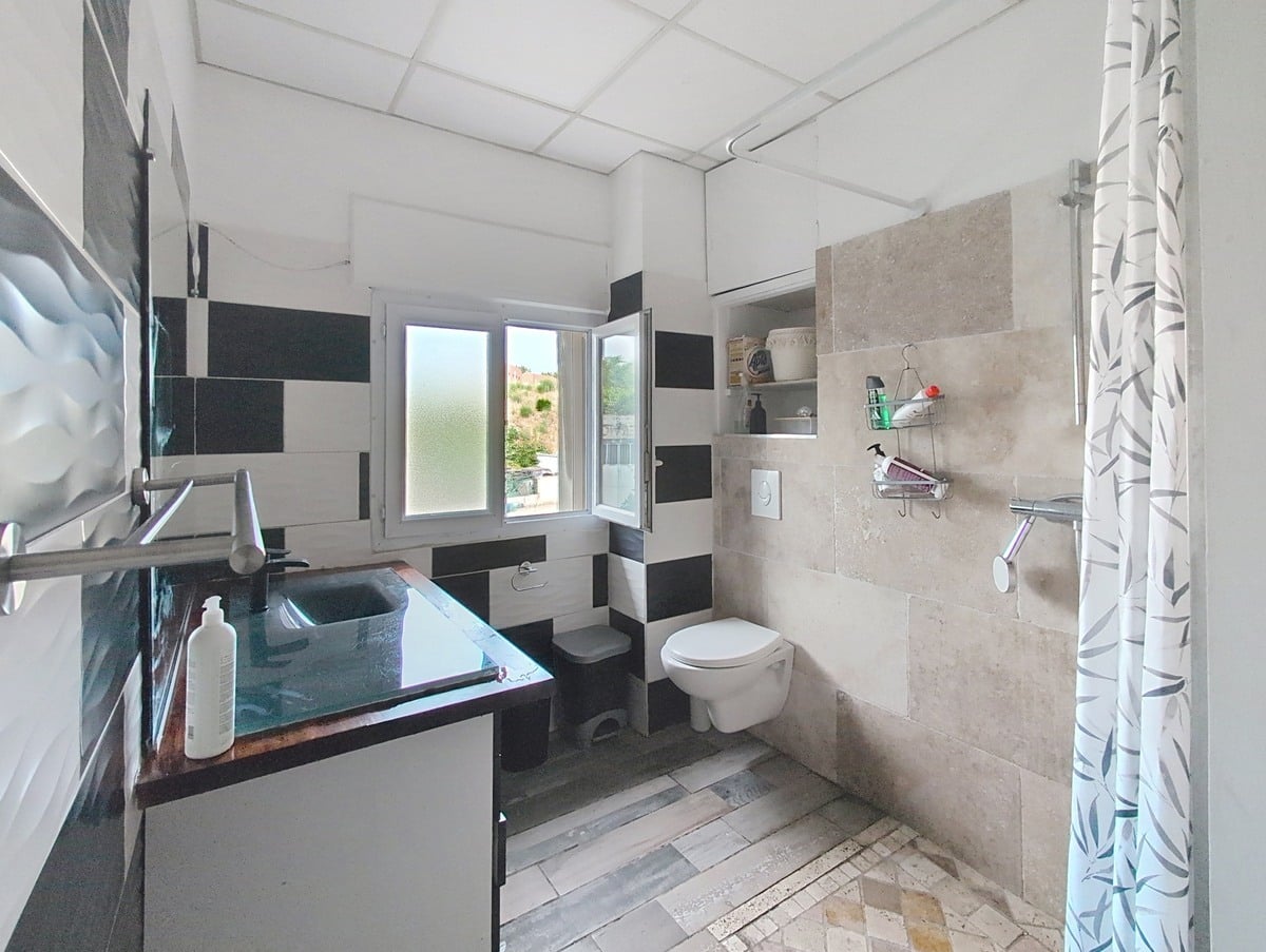 Salle d'eau avec carrelage mural en damier noir et blanc, sol en dalles imitation bois gris et ancien carrelage en terre cuite, équipée d'un WC suspendu blanc, d'une douche à l'italienne avec rideau à motifs végétaux et d'un meuble vasque noir à plan de travail sombre. Une fenêtre à battants apporte de la lumière naturelle et donne vue sur des arbres.