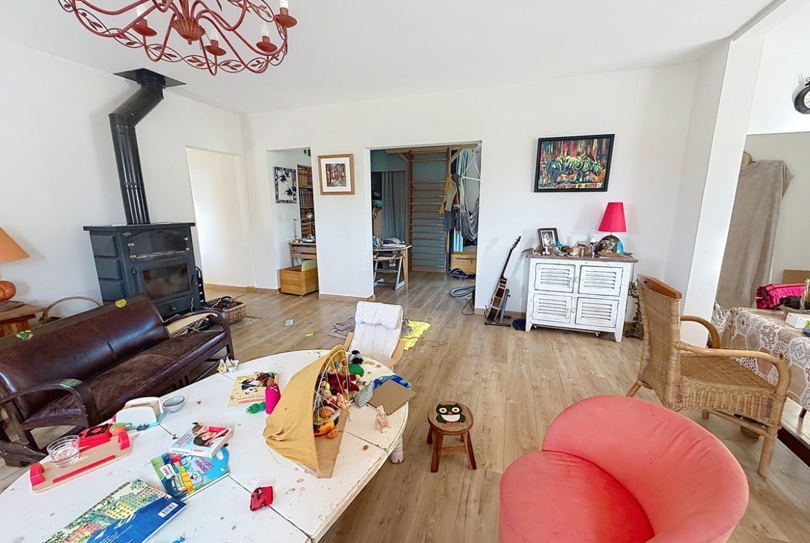 Salon aux murs blancs avec sol en parquet stratifié clair, équipé d'un poêle à bois en fonte noire avec conduit apparent et d'un canapé en cuir marron foncé. La pièce est meublée de plusieurs sièges dont un fauteuil rouge et un fauteuil rotin, avec un lustre en métal forgé rouge en suspension au plafond et des ouvertures vers un couloir avec escalier visible en arrière-plan.