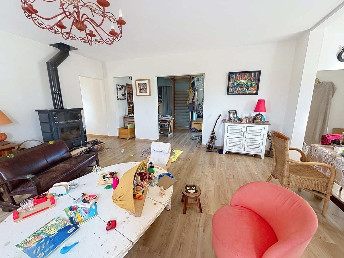 Salon aux murs blancs avec sol en parquet stratifié clair, équipé d'un poêle à bois en fonte noire avec conduit apparent et d'un canapé en cuir marron foncé. La pièce est meublée de plusieurs sièges dont un fauteuil rouge et un fauteuil rotin, avec un lustre en métal forgé rouge en suspension au plafond et des ouvertures vers un couloir avec escalier visible en arrière-plan.