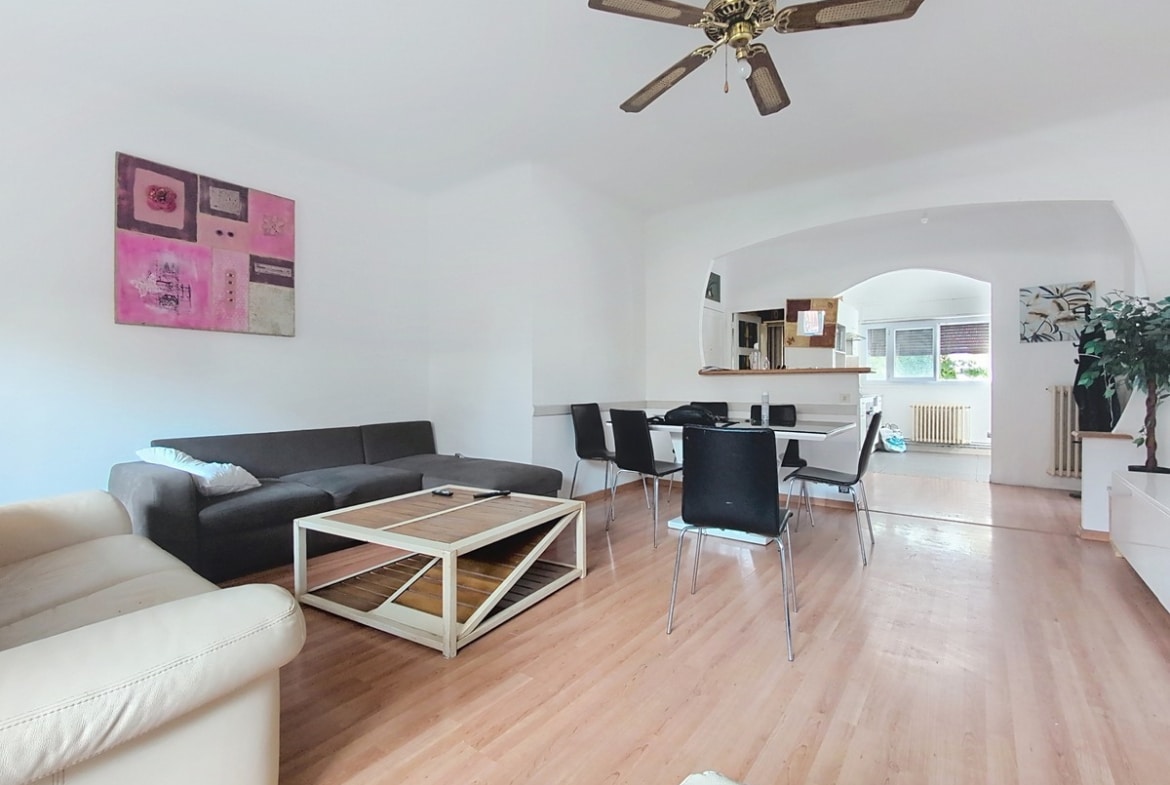 Séjour avec sol en parquet stratifié clair, plafond blanc à voûte et ventilateur en rotin, comprenant un canapé d'angle gris anthracite, une table basse en bois naturel et une table de salle à manger noire entourée de six chaises noires. Une arche blanche en fond de pièce ouvre sur la cuisine et l'entrée, avec une toile abstraite rose et blanche accrochée au mur.