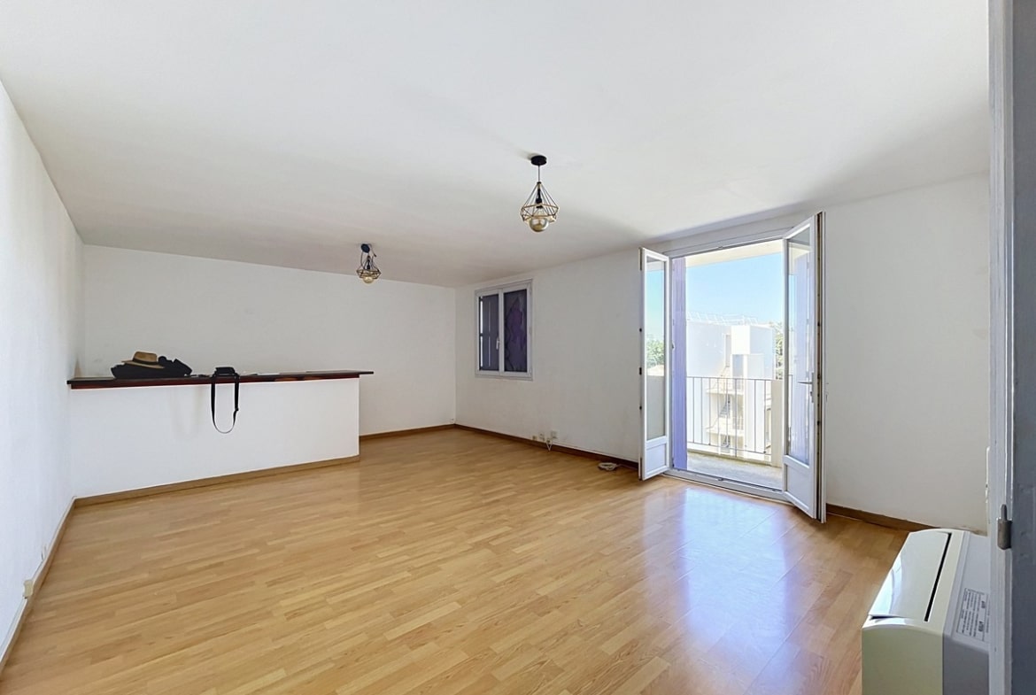 Grande pièce de vie lumineuse aux murs blancs avec sol en parquet stratifié clair, dotée d'une double porte-fenêtre ouverte donnant directement sur un balcon avec vue sur des immeubles résidentiels et ciel bleu. Une étagère murale en bois sombre court le long du mur gauche, et une suspension géométrique en métal noir est fixée au plafond blanc. Un système de climatisation mural est visible en bas à droite.