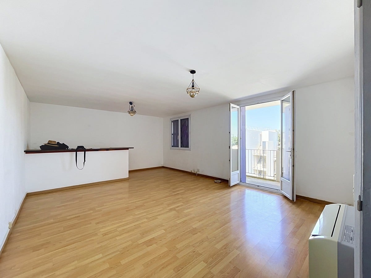 Grande pièce de vie lumineuse aux murs blancs avec sol en parquet stratifié clair, dotée d'une double porte-fenêtre ouverte donnant directement sur un balcon avec vue sur des immeubles résidentiels et ciel bleu. Une étagère murale en bois sombre court le long du mur gauche, et une suspension géométrique en métal noir est fixée au plafond blanc. Un système de climatisation mural est visible en bas à droite.