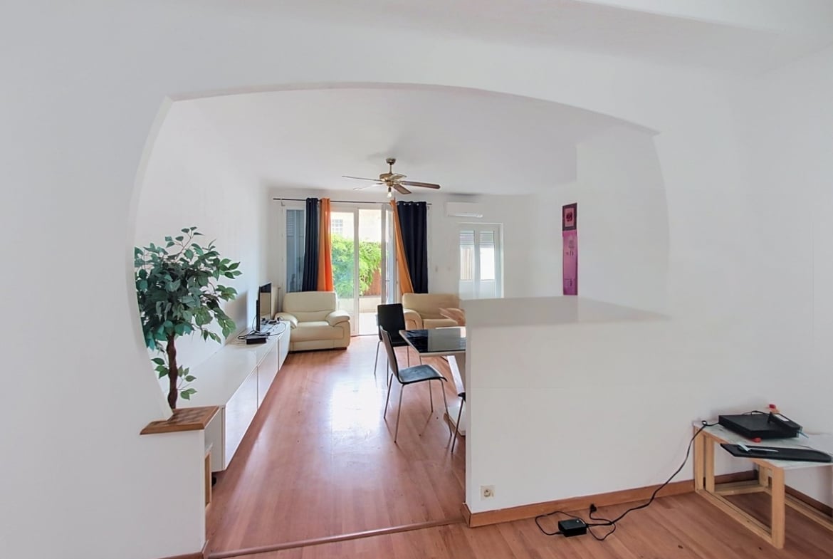 Pièce de vie avec sol en parquet clair, plafond voûté peint en blanc et ventilateur de plafond en laiton, ouvrant sur un salon meublé de deux fauteuils crème et d'une table de travail noire. Un bar de séparation en blanc délimite l'espace cuisine du séjour, avec vue sur un extérieur végétalisé à travers des rideaux noirs et orange.