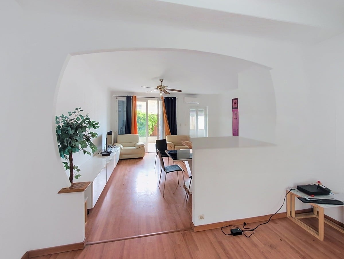 Pièce de vie avec sol en parquet clair, plafond voûté peint en blanc et ventilateur de plafond en laiton, ouvrant sur un salon meublé de deux fauteuils crème et d'une table de travail noire. Un bar de séparation en blanc délimite l'espace cuisine du séjour, avec vue sur un extérieur végétalisé à travers des rideaux noirs et orange.
