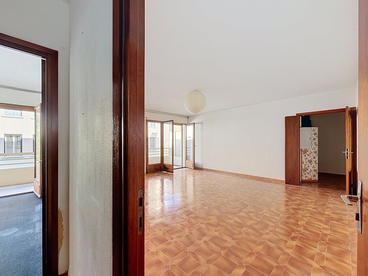 Vue depuis l'entrée du séjour au sol en carrelage tomettes hexagonales terre cuite, avec des portes-fenêtres en bois en fond donnant sur la terrasse extérieure. Une porte en bois sombre à droite donne accès à une pièce adjacente.