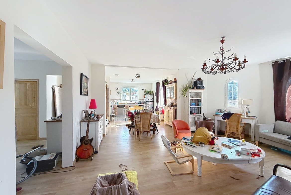 Grande pièce de vie à plan ouvert avec sol en parquet stratifié clair, comprenant un salon avec canapé gris et table basse blanche ovale, communiquant directement avec la salle à manger et la cuisine en arrière-plan. Un lustre en métal forgé noir et une fenêtre cintrée avec rideau bordeaux apportent du caractère à la pièce lumineuse aux murs blancs.