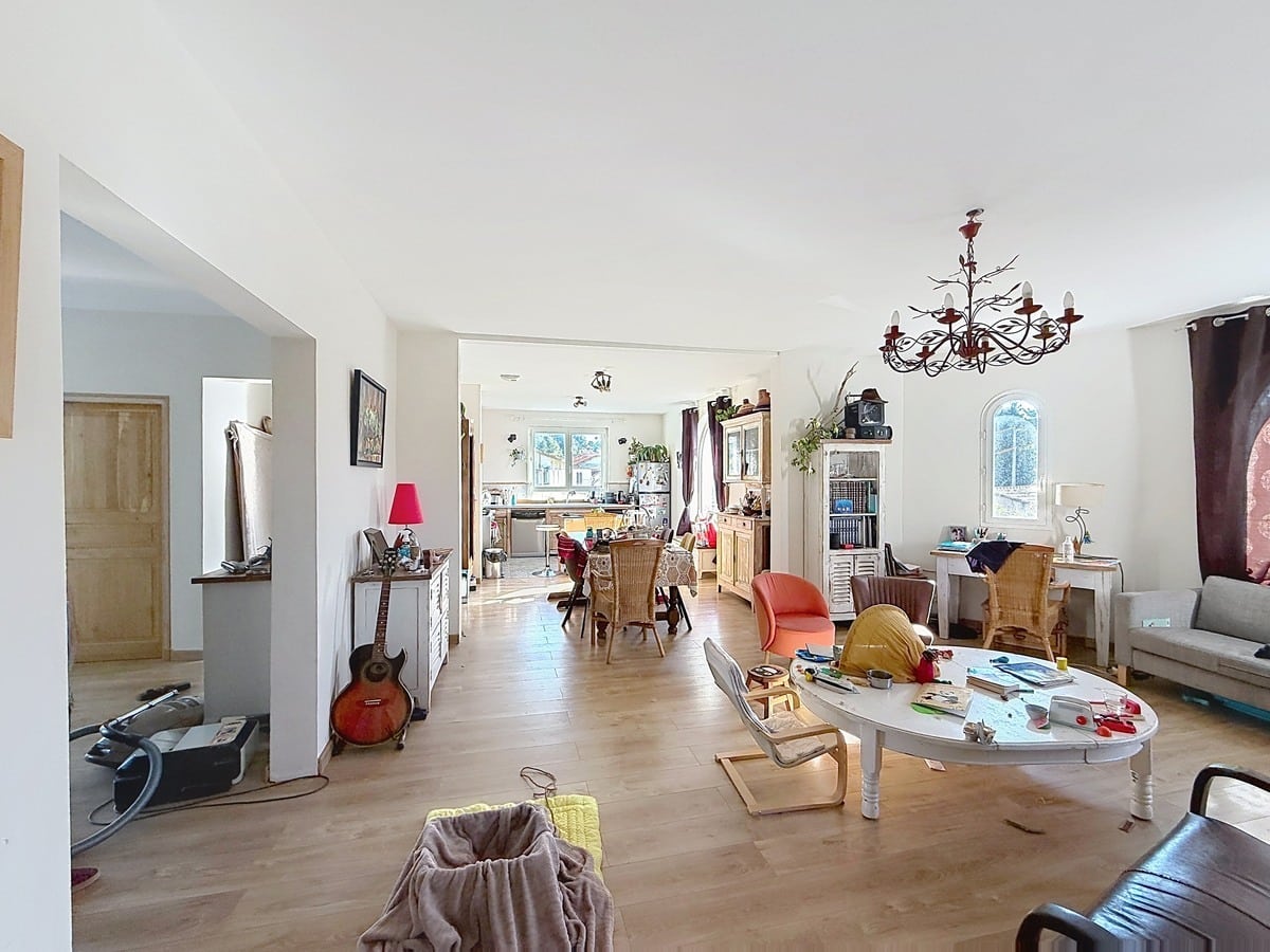 Grande pièce de vie à plan ouvert avec sol en parquet stratifié clair, comprenant un salon avec canapé gris et table basse blanche ovale, communiquant directement avec la salle à manger et la cuisine en arrière-plan. Un lustre en métal forgé noir et une fenêtre cintrée avec rideau bordeaux apportent du caractère à la pièce lumineuse aux murs blancs.