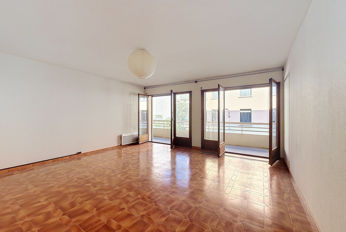Grand séjour vide au sol en carrelage tomettes hexagonales de couleur terre cuite, avec trois portes-fenêtres en bois donnant sur une terrasse filante. Le plafond blanc est éclairé par un luminaire boule en papier blanc.