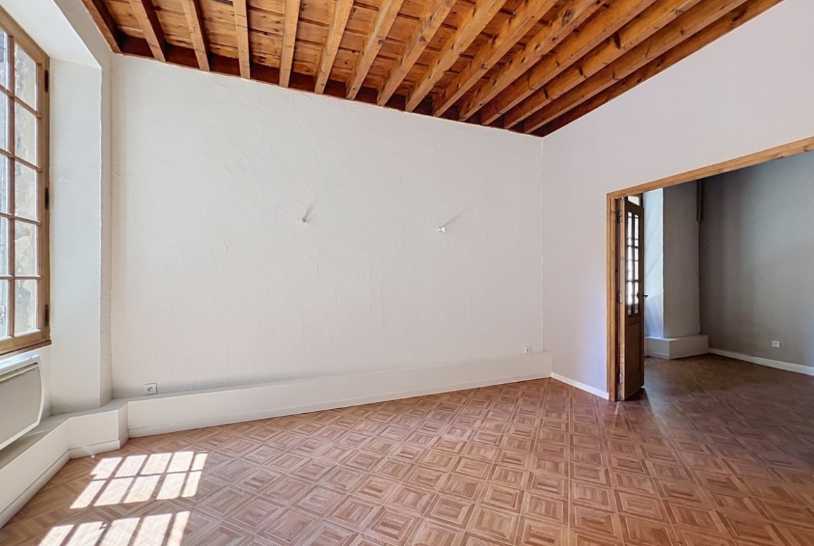 Pièce de séjour vide avec plafond à poutres en bois apparentes et murs blancs. Sol en parquet point de Hongrie et grande fenêtre à petits carreaux encadrée de bois laissant entrer la lumière naturelle.