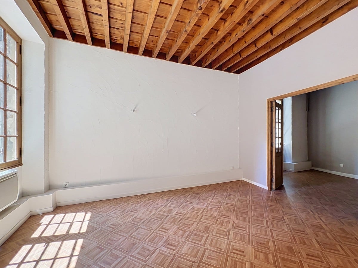 Pièce de séjour vide avec plafond à poutres en bois apparentes et murs blancs. Sol en parquet point de Hongrie et grande fenêtre à petits carreaux encadrée de bois laissant entrer la lumière naturelle.