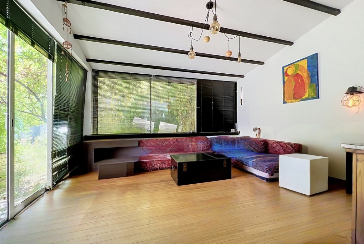 Salon avec plafond à poutres apparentes peintes en noir, équipé d'un grand canapé d'angle modulable en velours rouge et violet posé face à une baie vitrée coulissante donnant sur un espace végétalisé. Le sol est en parquet bambou clair, un lustre composé de plusieurs ampoules à filament est suspendu au plafond.