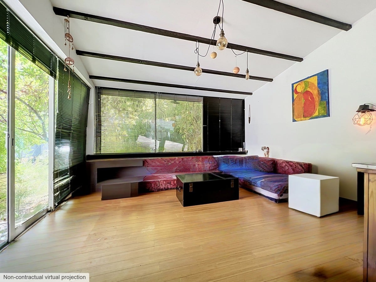 Salon avec plafond à poutres apparentes peintes en noir, équipé d'un grand canapé d'angle modulable en velours rouge et violet posé face à une baie vitrée coulissante donnant sur un espace végétalisé. Le sol est en parquet bambou clair, un lustre composé de plusieurs ampoules à filament est suspendu au plafond.