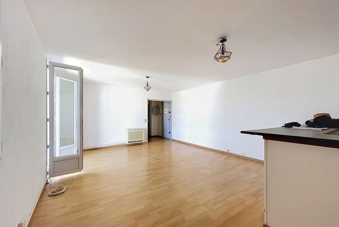 Pièce de vie rectangulaire aux murs blancs avec sol en parquet stratifié clair, éclairée par deux suspensions design en métal doré géométrique. Une porte-fenêtre avec cadre gris donne sur l'extérieur à gauche, tandis qu'un radiateur mural blanc est positionné sous l'entrée de la pièce visible au fond. Un comptoir de cuisine sombre apparaît en amorce sur la droite.