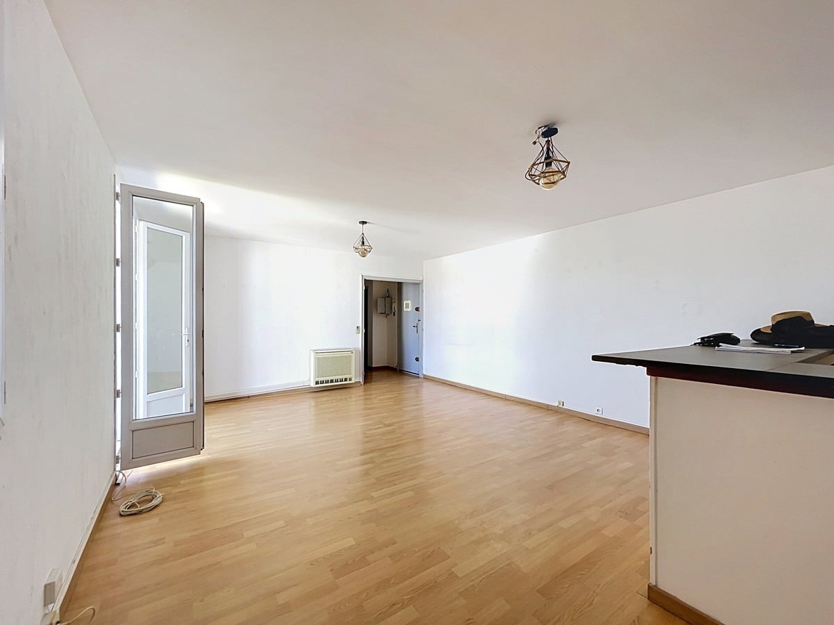 Pièce de vie rectangulaire aux murs blancs avec sol en parquet stratifié clair, éclairée par deux suspensions design en métal doré géométrique. Une porte-fenêtre avec cadre gris donne sur l'extérieur à gauche, tandis qu'un radiateur mural blanc est positionné sous l'entrée de la pièce visible au fond. Un comptoir de cuisine sombre apparaît en amorce sur la droite.