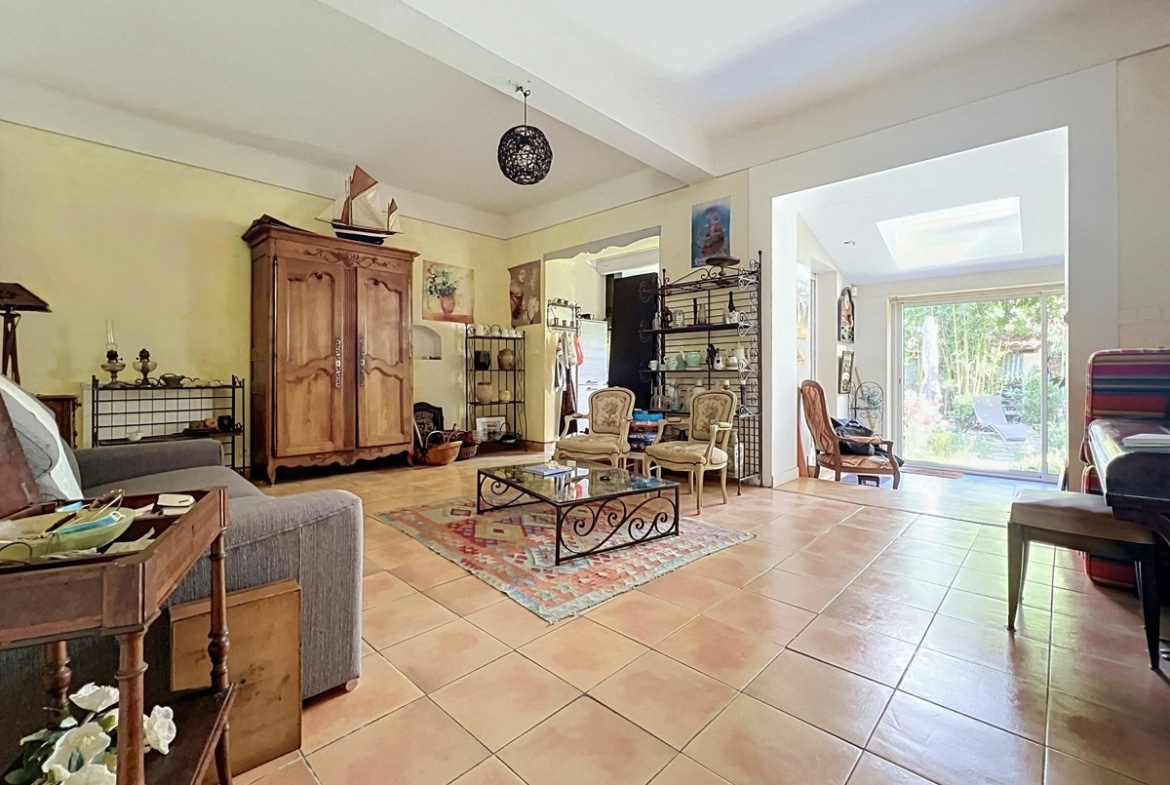 Grand salon avec sol en carrelage terre cuite, armoire normande en chêne massif et table basse en fer forgé sur tapis kilim multicolore. Deux canapés gris, plusieurs fauteuils et ouverture en arc vers une véranda lumineuse donnant sur le jardin.