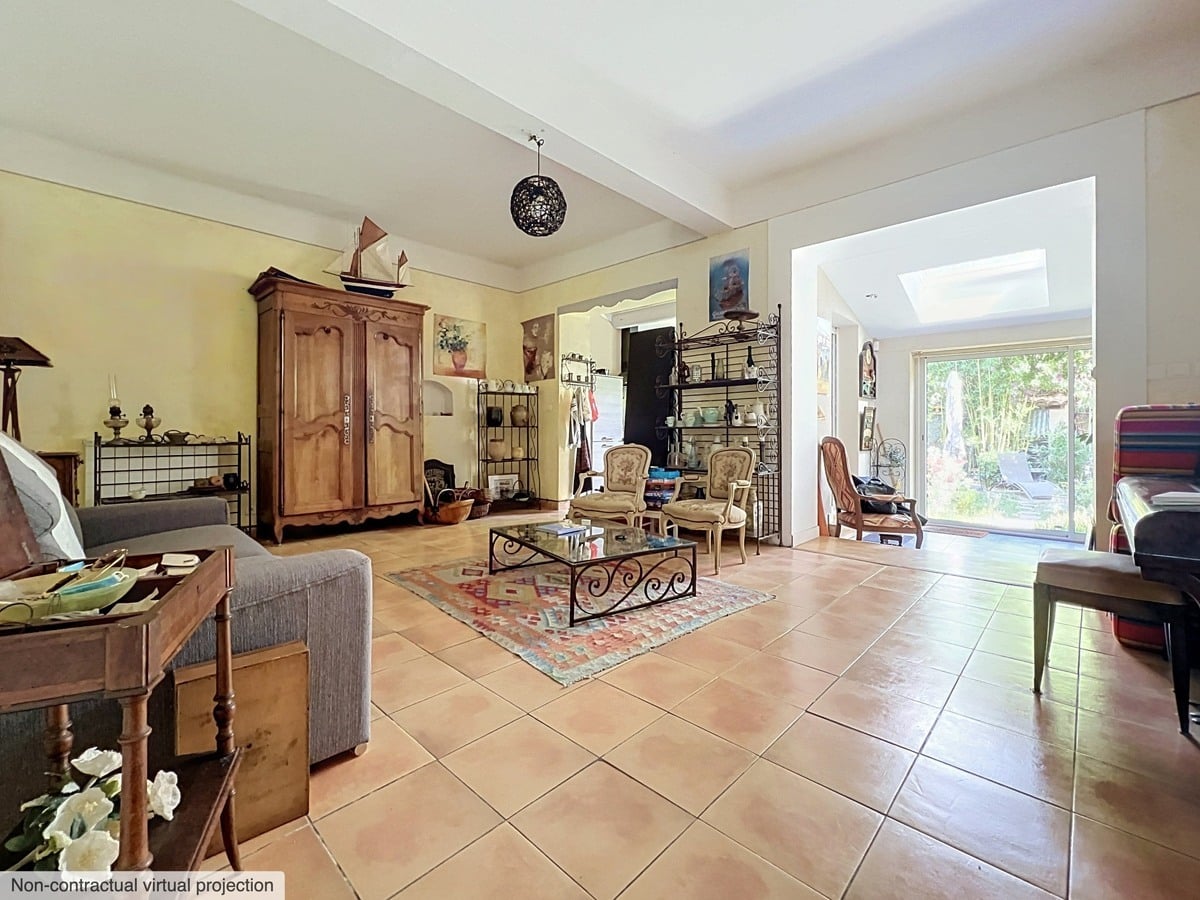Grand salon avec sol en carrelage terre cuite, armoire normande en chêne massif et table basse en fer forgé sur tapis kilim multicolore. Deux canapés gris, plusieurs fauteuils et ouverture en arc vers une véranda lumineuse donnant sur le jardin.