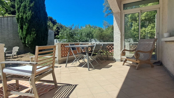 Terrasse couverte carrelée de dalles beige orangé, équipée d'une table de jardin grise avec chaises pliantes et de fauteuils en bois ancien, délimitée par un muret en briques apparentes. En fond, un jardin avec cyprès et arbres feuillus sous un ciel bleu intense, avec accès à la maison par une baie vitrée coulissante.