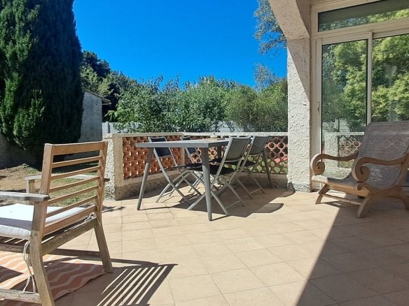 Terrasse couverte carrelée de dalles beige orangé, équipée d'une table de jardin grise avec chaises pliantes et de fauteuils en bois ancien, délimitée par un muret en briques apparentes. En fond, un jardin avec cyprès et arbres feuillus sous un ciel bleu intense, avec accès à la maison par une baie vitrée coulissante.