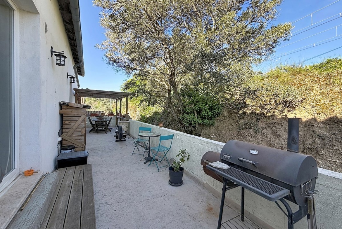 Terrasse en béton longeant la façade blanche de la maison, équipée d'un barbecue en métal noir, de chaises pliantes bleues et d'une table ronde en mosaïque. Un olivier mature domine l'arrière-plan au-dessus d'un muret blanc, avec une pergola en bois visible en fond de terrasse.