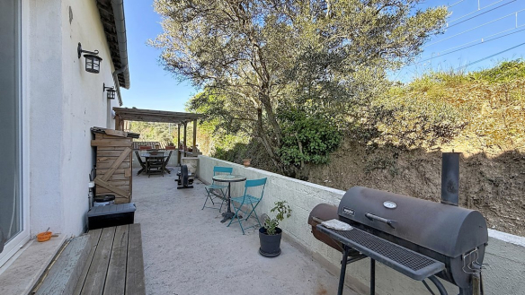 Terrasse en béton longeant la façade blanche de la maison, équipée d'un barbecue en métal noir, de chaises pliantes bleues et d'une table ronde en mosaïque. Un olivier mature domine l'arrière-plan au-dessus d'un muret blanc, avec une pergola en bois visible en fond de terrasse.