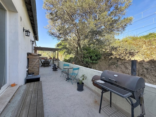 Terrasse en béton longeant la façade blanche de la maison, équipée d'un barbecue en métal noir, de chaises pliantes bleues et d'une table ronde en mosaïque. Un olivier mature domine l'arrière-plan au-dessus d'un muret blanc, avec une pergola en bois visible en fond de terrasse.