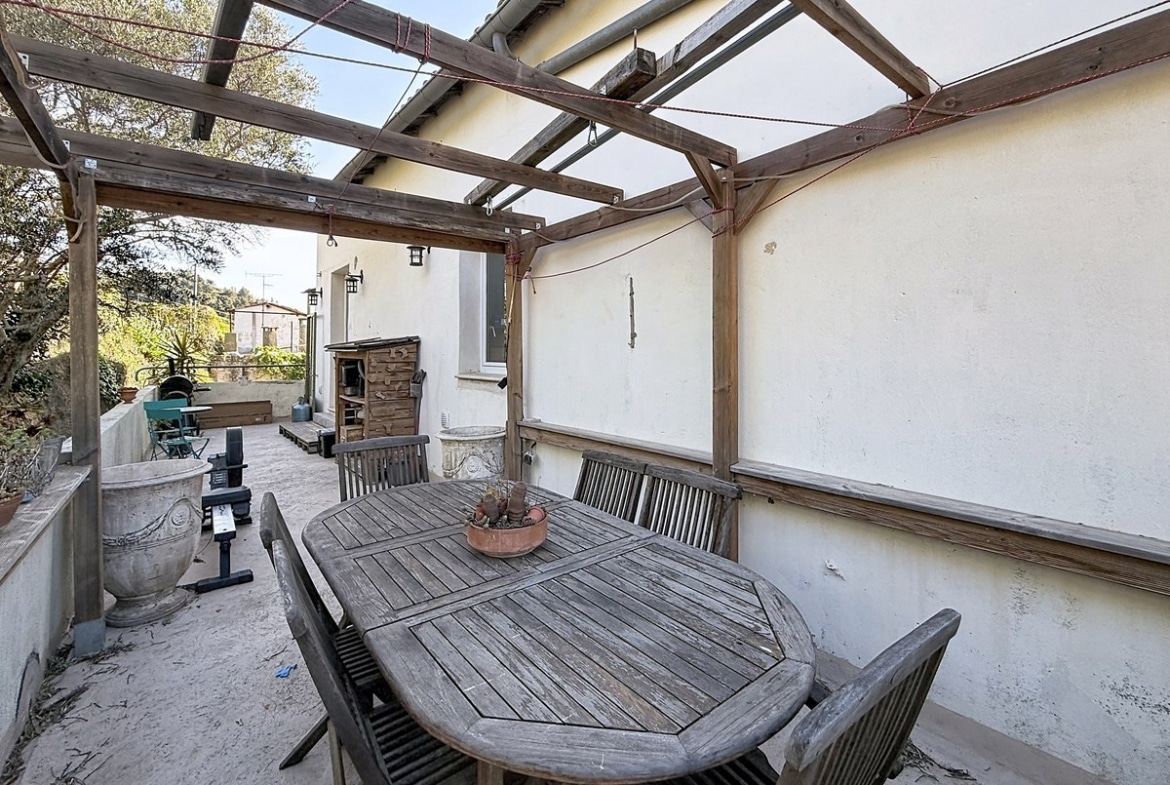 Terrasse extérieure couverte par une pergola en bois grisé avec structure ouverte et cordes rouges tendues, sous laquelle est installée une table de jardin ovale en teck avec six chaises assorties. En fond, une deuxième zone de terrasse accueille du mobilier de jardin supplémentaire et un four à pizza en terre cuite contre le mur de la maison.