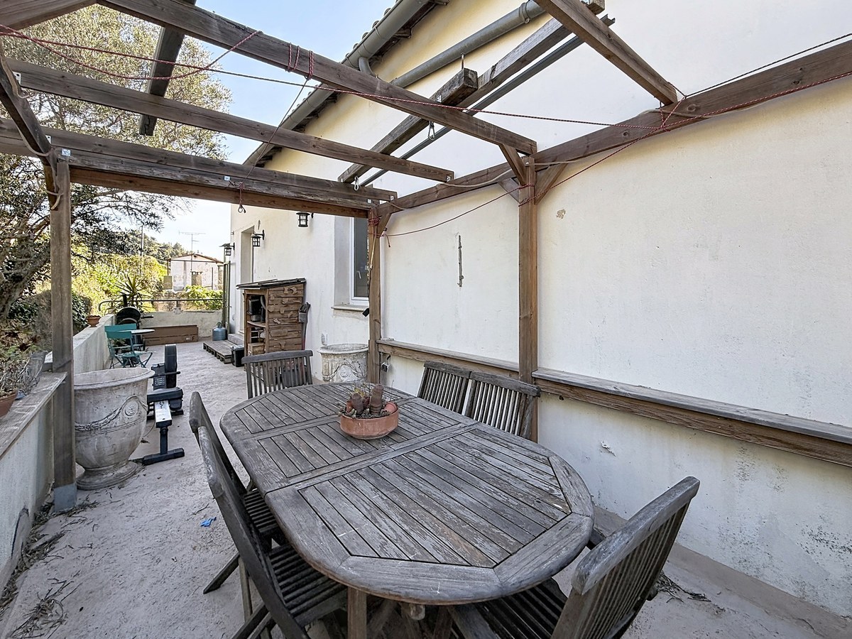 Terrasse extérieure couverte par une pergola en bois grisé avec structure ouverte et cordes rouges tendues, sous laquelle est installée une table de jardin ovale en teck avec six chaises assorties. En fond, une deuxième zone de terrasse accueille du mobilier de jardin supplémentaire et un four à pizza en terre cuite contre le mur de la maison.