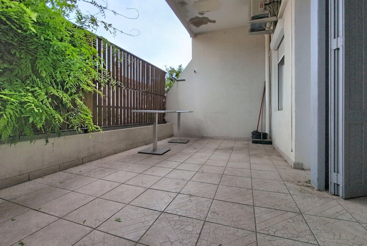 Terrasse carrelée de dalles beiges craquelées, délimitée par un muret blanc et un panneau brise-vue en lattes de bois brun, avec un mur végétalisé de fougères vertes sur la gauche. Une unité extérieure de climatisation est fixée en hauteur sur la façade blanche, et deux tables de bistrot en métal gris sont disposées sur l'espace extérieur.