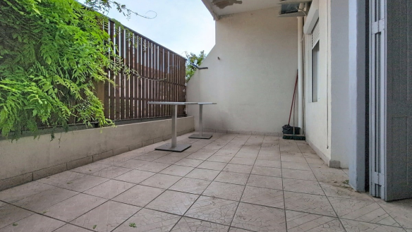 Terrasse carrelée de dalles beiges craquelées, délimitée par un muret blanc et un panneau brise-vue en lattes de bois brun, avec un mur végétalisé de fougères vertes sur la gauche. Une unité extérieure de climatisation est fixée en hauteur sur la façade blanche, et deux tables de bistrot en métal gris sont disposées sur l'espace extérieur.